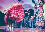 A flamenco dancer performs energetically, with a vibrant red ruffled dress swirling in motion. Musicians, including a guitarist, accompany with lively music in a traditional setting. The room has arched walls and the audience is engaged in the performance.