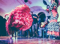 A flamenco dancer performs energetically, with a vibrant red ruffled dress swirling in motion. Musicians, including a guitarist, accompany with lively music in a traditional setting. The room has arched walls and the audience is engaged in the performance.