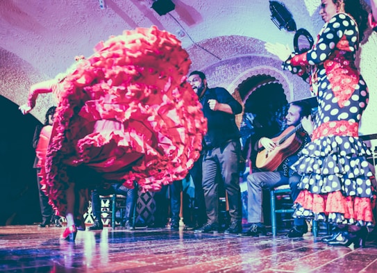 A flamenco dancer performs energetically, with a vibrant red ruffled dress swirling in motion. Musicians, including a guitarist, accompany with lively music in a traditional setting. The room has arched walls and the audience is engaged in the performance.