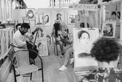 An art studio filled with several people engaged in drawing or painting portraits. Easels with various portrait sketches and paintings are scattered throughout the room. The atmosphere is busy and artistic, with numerous portraits displayed on the walls and on easels.