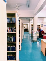 A library interior with rows of book shelves filled with books, a blue floor, ceiling fans, and chairs and desks arranged for reading. A stand with magazines and a clean, organized ambiance can also be seen.