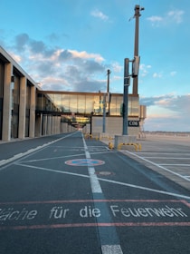 A modern airport terminal with a glass-walled walkway connecting the buildings, located next to an empty tarmac. The sky is filled with light clouds illuminated by the setting or rising sun, creating a serene atmosphere. The word 'C06' is visible on a sign, and there is a designated path painted on the ground with text in German.