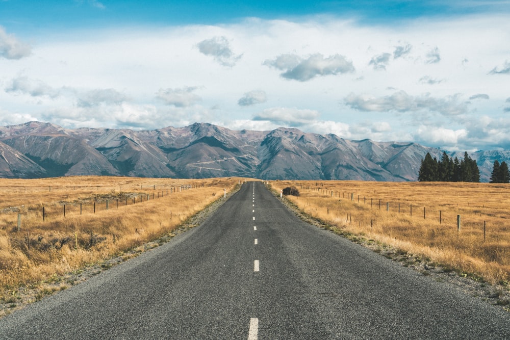 gray concrete road between brown grass field near mountain under white clouds and blue sky during