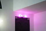 A doorway labeled as a toilet is illuminated with pink lighting, set against a white wall. The door is dark-colored with a specific geometric pattern.