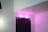 A doorway labeled as a toilet is illuminated with pink lighting, set against a white wall. The door is dark-colored with a specific geometric pattern.