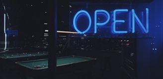 A dimly lit room is visible through glass, featuring a neon 'OPEN' sign glowing in bright blue. Inside, a pool table with scattered balls is partially visible, surrounded by a low-lit, cozy atmosphere.