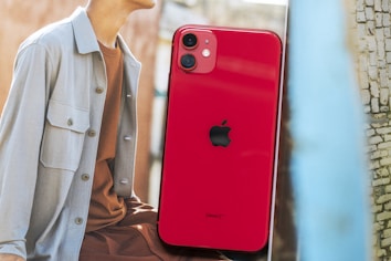 A red smartphone with a dual-camera setup and a prominent logo is positioned in the foreground. In the background, part of a person wearing casual attire is visible, with a focus on the upper torso dressed in a light-colored jacket over a brown shirt.