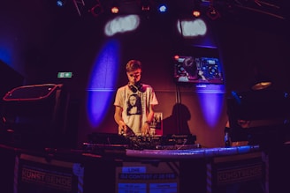 A DJ is performing at an event, standing behind a mixing console with headphones around his neck. The room is dimly lit with colorful spotlights, creating vibrant red, blue, and purple hues. A screen behind the DJ displays a live feed of the equipment he is using.