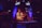 A DJ is performing at an event, standing behind a mixing console with headphones around his neck. The room is dimly lit with colorful spotlights, creating vibrant red, blue, and purple hues. A screen behind the DJ displays a live feed of the equipment he is using.