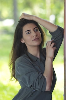 A person with long hair and a relaxed expression is gently posing with one hand resting on their head and the other near their face. They are wearing a casual, long-sleeved shirt. The background features a soft, blurred natural setting with varying shades of green suggesting an outdoor environment.