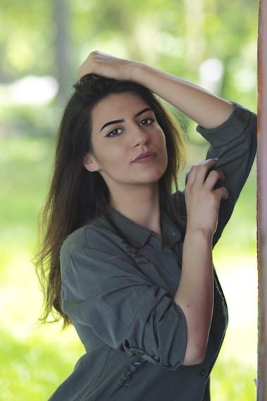 A person with long hair and a relaxed expression is gently posing with one hand resting on their head and the other near their face. They are wearing a casual, long-sleeved shirt. The background features a soft, blurred natural setting with varying shades of green suggesting an outdoor environment.