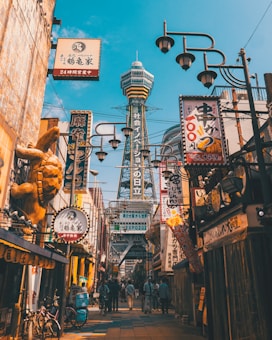 A bustling urban street features various shop signs with Japanese characters, colorful decorations, and a prominent tower with a unique design in the background. People walk along the street, creating a lively atmosphere, and bicycles are parked along the sides.