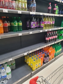 A supermarket aisle displays various bottles of beverages, including sodas and water, arranged on shelves. The shelves contain bottles of different colors, such as green, yellow, orange, and clear. A shopping cart is visible in the foreground, partially empty, with a red handle.