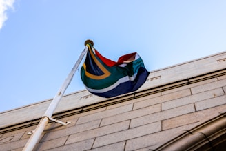 A flag with vibrant colors waves in the wind, mounted on a pole attached to a stone building. The flag's design features a combination of green, yellow, black, white, red, and blue shades. The pole is securely fastened to the building, which has a textured stone facade. The sky in the background is clear and bright blue.