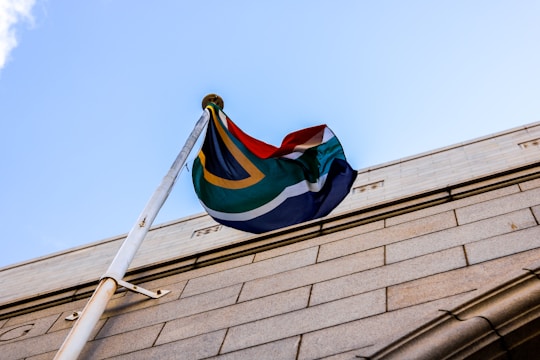 A flag with vibrant colors waves in the wind, mounted on a pole attached to a stone building. The flag's design features a combination of green, yellow, black, white, red, and blue shades. The pole is securely fastened to the building, which has a textured stone facade. The sky in the background is clear and bright blue.