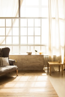 A cozy living room bathed in warm, natural light streaming through large, sheer curtains. A classic-style sofa with a striped cushion is on one side, and a simple wooden table holds a small candle. The wooden floor adds to the rustic charm, and a windowsill hosts a few small decorative items, including a plant and a ceramic object.