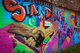 A colorful mural on a brick wall featuring large text in bold graffiti style reading 'Stand with Us'. The artwork includes different flags, a crescent moon, and a star, along with a diverse range of patterns and symbols. There are two hands shaking prominently in the center, emphasizing unity and solidarity.