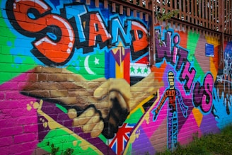 A colorful mural on a brick wall featuring large text in bold graffiti style reading 'Stand with Us'. The artwork includes different flags, a crescent moon, and a star, along with a diverse range of patterns and symbols. There are two hands shaking prominently in the center, emphasizing unity and solidarity.