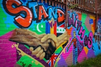 A colorful mural on a brick wall featuring large text in bold graffiti style reading 'Stand with Us'. The artwork includes different flags, a crescent moon, and a star, along with a diverse range of patterns and symbols. There are two hands shaking prominently in the center, emphasizing unity and solidarity.