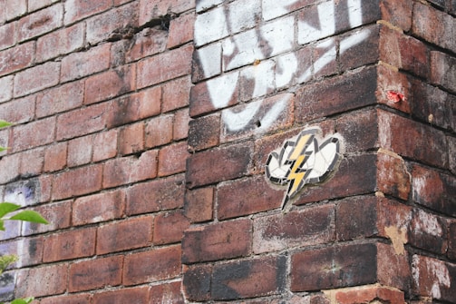 A section of a brick wall with graffiti and a sticker of a lightning bolt. The bricks are various shades of red and brown, and the wall has white spray-painted markings. A small red and white label is affixed to the corner of the wall.