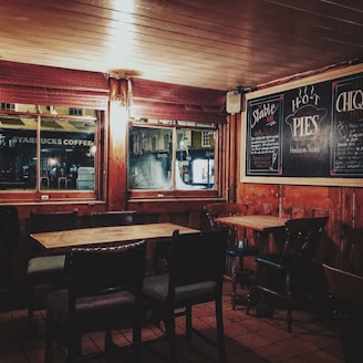 A cozy, dimly lit pub interior featuring wooden tables and chairs arranged orderly. Chalkboards on the walls advertise hot pies. Large windows provide a glimpse of the street outside, with signs from nearby shops visible.