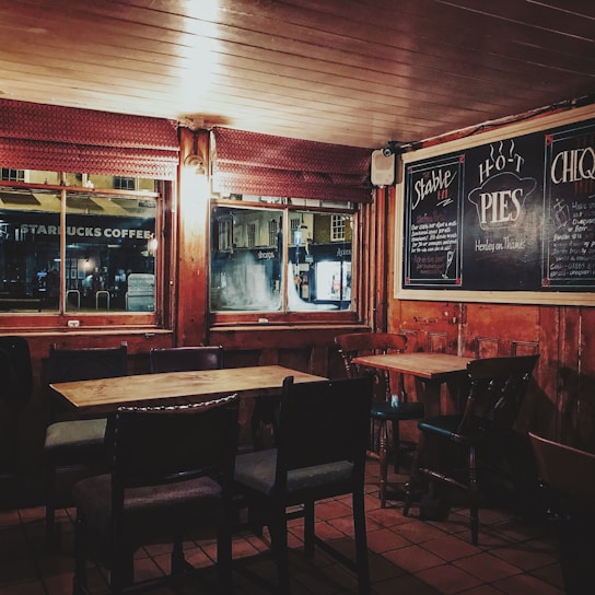 A cozy, dimly lit pub interior featuring wooden tables and chairs arranged orderly. Chalkboards on the walls advertise hot pies. Large windows provide a glimpse of the street outside, with signs from nearby shops visible.