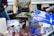 People are engaged in a painting activity, holding canvases with abstract designs over a table covered with a plastic sheet. Various cups filled with paint, paper towels, and other painting supplies are scattered on the table. They are wearing aprons and gloves to protect themselves from paint.
