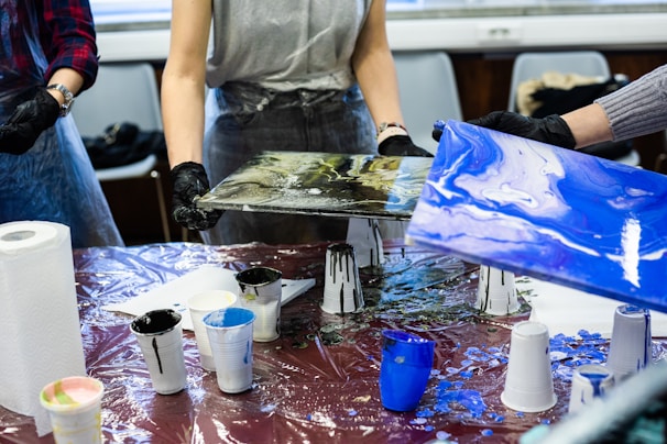 People are engaged in a painting activity, holding canvases with abstract designs over a table covered with a plastic sheet. Various cups filled with paint, paper towels, and other painting supplies are scattered on the table. They are wearing aprons and gloves to protect themselves from paint.