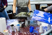 People are engaged in a painting activity, holding canvases with abstract designs over a table covered with a plastic sheet. Various cups filled with paint, paper towels, and other painting supplies are scattered on the table. They are wearing aprons and gloves to protect themselves from paint.