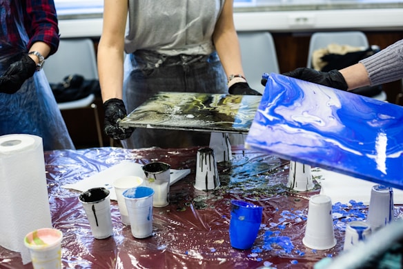 People are engaged in a painting activity, holding canvases with abstract designs over a table covered with a plastic sheet. Various cups filled with paint, paper towels, and other painting supplies are scattered on the table. They are wearing aprons and gloves to protect themselves from paint.