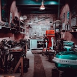 A vintage workshop filled with various tools and machinery. Shelves line the walls, holding different equipment and small items. A wooden door at the back is adorned with hanging tools, and the floor is lined with a metal grate. The lighting is dim, creating a rustic and antique atmosphere.