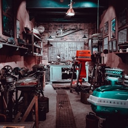A vintage workshop filled with various tools and machinery. Shelves line the walls, holding different equipment and small items. A wooden door at the back is adorned with hanging tools, and the floor is lined with a metal grate. The lighting is dim, creating a rustic and antique atmosphere.