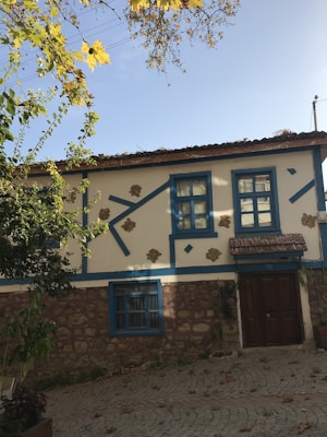A quaint house with stone and white plaster walls, accented with blue wooden beams and window frames. The roof is made of dark tiles and there is a large wooden door at the entrance. Leaves and branches from nearby trees frame the top of the image, casting shadows on the house.