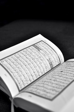 An open book with text written in Arabic script is prominently displayed. The pages are lightly tinted in sepia tones, suggesting an old or sacred text. The background is dark, highlighting the text.