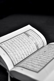 An open book with text written in Arabic script is prominently displayed. The pages are lightly tinted in sepia tones, suggesting an old or sacred text. The background is dark, highlighting the text.