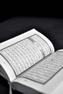 An open book with text written in Arabic script is prominently displayed. The pages are lightly tinted in sepia tones, suggesting an old or sacred text. The background is dark, highlighting the text.
