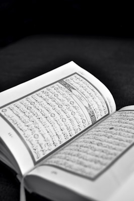 An open book with text written in Arabic script is prominently displayed. The pages are lightly tinted in sepia tones, suggesting an old or sacred text. The background is dark, highlighting the text.