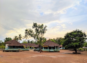 A rural setting featuring small buildings with tiled roofs surrounded by trees under a partly cloudy sky. The area is open with a dirt ground and sparse vegetation, creating a peaceful rural atmosphere.