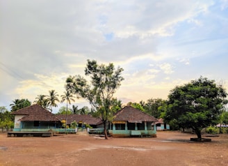A rural setting featuring small buildings with tiled roofs surrounded by trees under a partly cloudy sky. The area is open with a dirt ground and sparse vegetation, creating a peaceful rural atmosphere.