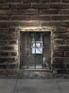 A brick wall with a rectangular opening in the center, containing a gas meter. The meter is enclosed behind a metal grate. The bricks are various shades of brown and red, and the area around the opening shows signs of wear and age.