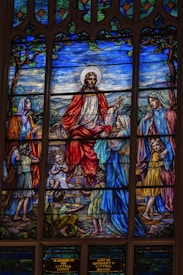 A stained glass window depicting a biblical scene with central figure in a red and white robe surrounded by several other figures, including children and adults. The background features a landscape with trees and a blue sky.