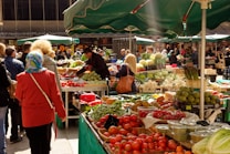 A bustling outdoor market with numerous stalls displaying a variety of fresh produce including tomatoes, artichokes, lettuces, and other vegetables. People are browsing and shopping, with some inspecting the produce closely. The stalls are covered with green umbrellas providing shade.