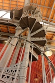 An intricate and ornate spiral staircase made of metal, featuring detailed patterns and a robust structure. It ascends elegantly under a ceiling with a wooden grid pattern. The setting is enhanced by large windows and industrial-style lighting.