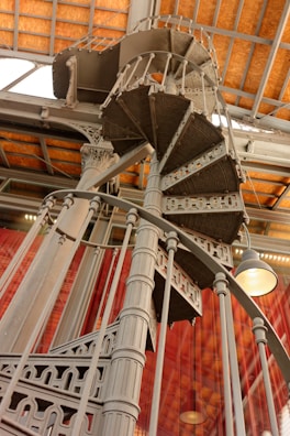 An intricate and ornate spiral staircase made of metal, featuring detailed patterns and a robust structure. It ascends elegantly under a ceiling with a wooden grid pattern. The setting is enhanced by large windows and industrial-style lighting.