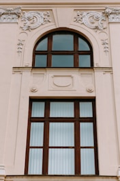 A large, ornate window set in a beige-colored building wall features intricate decorative moldings. The window has a wooden frame with multiple panes, some of which are arched at the top. The embellishments surrounding the window include swirling floral patterns and relief designs.
