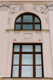 A large, ornate window set in a beige-colored building wall features intricate decorative moldings. The window has a wooden frame with multiple panes, some of which are arched at the top. The embellishments surrounding the window include swirling floral patterns and relief designs.