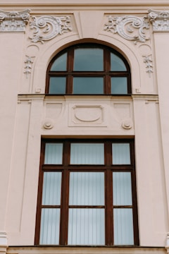 A large, ornate window set in a beige-colored building wall features intricate decorative moldings. The window has a wooden frame with multiple panes, some of which are arched at the top. The embellishments surrounding the window include swirling floral patterns and relief designs.