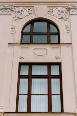 A large, ornate window set in a beige-colored building wall features intricate decorative moldings. The window has a wooden frame with multiple panes, some of which are arched at the top. The embellishments surrounding the window include swirling floral patterns and relief designs.