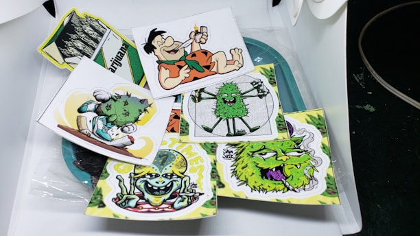 A collection of cartoonish and vibrant stickers featuring various humorous and playful characters, including a caveman with a cigar, smiling green creatures, and quirky designs. Each sticker is colorful and arranged in a casual, overlapping manner on a surface.
