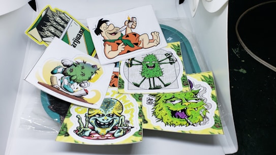 A collection of cartoonish and vibrant stickers featuring various humorous and playful characters, including a caveman with a cigar, smiling green creatures, and quirky designs. Each sticker is colorful and arranged in a casual, overlapping manner on a surface.